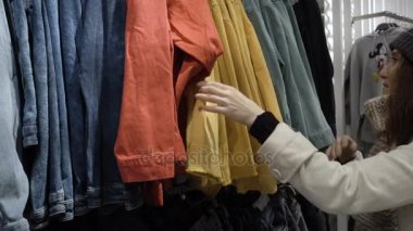 Elinde yeni giysiler, askı askı üzerinde farklı rahat renkli giysiler ile bakarak seçmek genç güzel kadın moda alışveriş merkezinde alışveriş yakın çekim