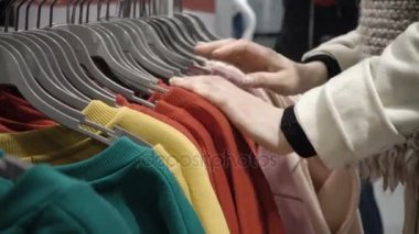 Elinde yeni giysiler, askı askı üzerinde farklı rahat renkli giysiler ile bakarak seçmek genç güzel kadın moda alışveriş merkezinde alışveriş yakın çekim