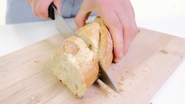 Mãos de mulher cortando pão na cozinha. Fechar as mãos femininas segurando faca, cortando a baguete branca na tábua de corte. Casa de culinária — Vídeo de Stock