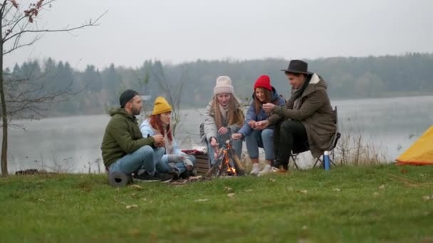 Skupina šťastných přátel kolem zapalování táboráku v lese pečení marshmallows mluvit a usmívat se. Turisté sedící kolem táborového ohně. Venkovní piknik v lese. — Stock video