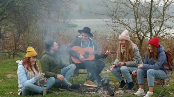 Happy friends sitting around a bonfire on the beach, playing guitar and singing — Stock video
