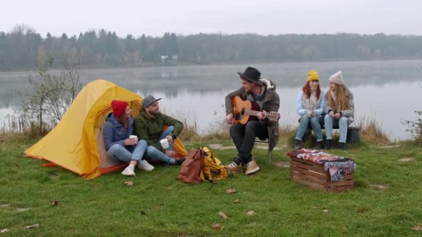 Ευτυχισμένοι φίλοι κάθονται γύρω από μια φωτιά στην παραλία, παίζοντας κιθάρα και τραγουδώντας — Αρχείο Βίντεο