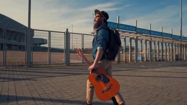 Hipster brodaty męski muzyk z ciemnymi, ciepłymi oczami spacerujący po parku w słoneczny dzień z gitarą — Wideo stockowe