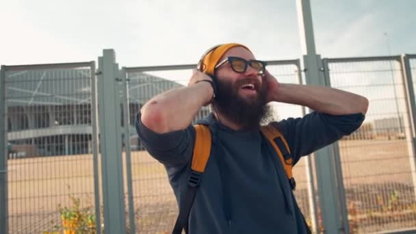 Ritratto ravvicinato di giovane hipster barbuto con cuffie ascoltare musica e sorridere alla strada della città — Video Stock