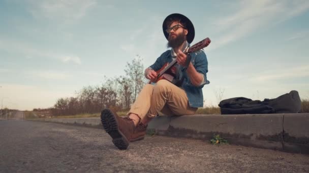 Молодой человек играет на гитаре и сидит на земле на городской дороге — стоковое видео