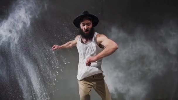 Beard guy a bboying style dancer does difficult tricks on the floor in a studio filled with flour on a black background. — Stock Video