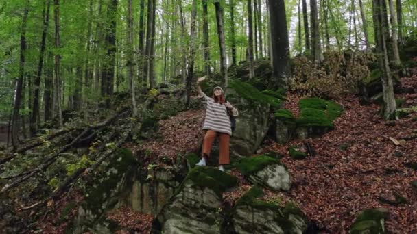 Giovane ragazza turistica felice sorridente in un cappello, con uno zaino grigio prende un selfie con il suo smartphone e monopiede, posa in un bellissimo luogo di foresta di montagna . — Video Stock