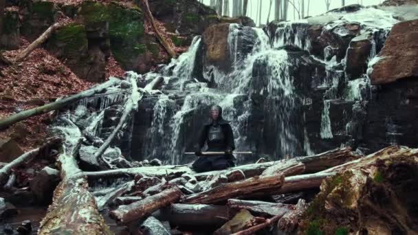 Człowiek praktykujący medytację mieczem bambusowym na tle wodospadu zimowego. — Wideo stockowe
