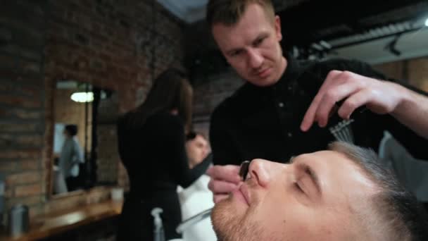 Um jovem bonito a rapar a barba na barbearia. Barba cortada com uma navalha reta em close-up. Barbeiro profissional barba barba do cliente em um salão com uma navalha reta à moda antiga — Vídeo de Stock