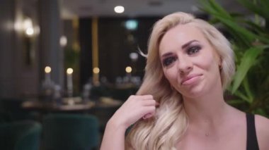 Akşam elbisesi giymiş sarışın bir kadın şık bir restoranın masasında oturan takım elbiseli bir adamla kadeh tokuşturuyor..