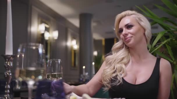 En blond kvinna i aftonklänning är klirrande glas med en man i kostym, sitter vid bordet i en fin restaurang. — Stockvideo