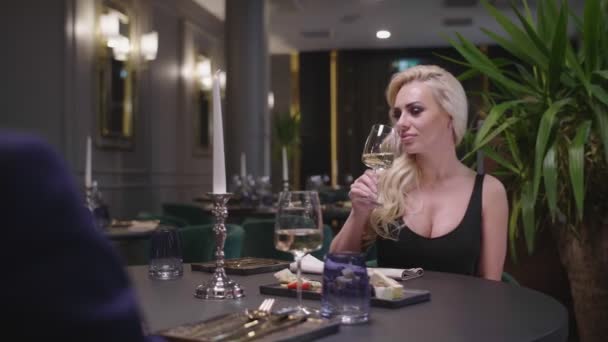 Seorang wanita pirang bergaun malam adalah berkacamata dengan seorang pria berjas, duduk di meja restoran. . — Stok Video