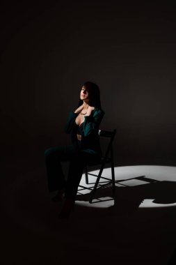 Alacakaranlıkta siyah sandalyede oturan güzel esmer kadının stüdyo fotoğrafı. Yüzüne bir ışık huzmesi vuruyor..