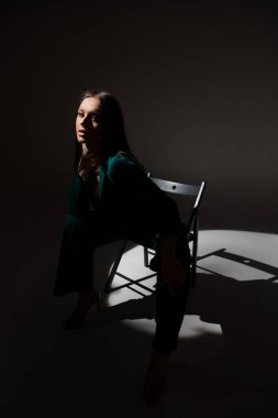 Alacakaranlıkta siyah sandalyede oturan güzel esmer kadının stüdyo fotoğrafı. Yüzüne bir ışık huzmesi vuruyor..