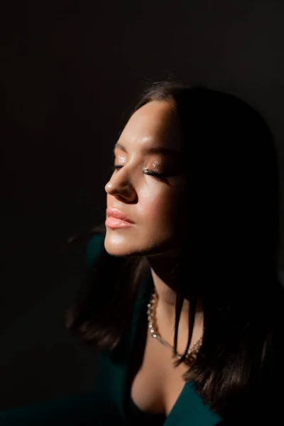 Studio portrait of pretty brunette woman in twilight sitting on black chair. A ray of light hits her face.