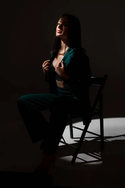 Studiofoto einer hübschen brünetten Frau in der Dämmerung, die auf einem schwarzen Stuhl sitzt. Ein Lichtstrahl trifft ihr Gesicht. — Stockfoto