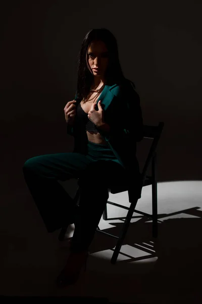 Studiová fotka krásné brunetky v soumraku sedící na černém křesle. Paprsek světla zasáhne její tvář. — Stock fotografie