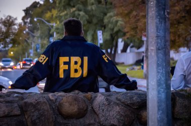 FBI logosu olan lacivert ceketli erkek bir FBI ajanı alacakaranlıkta arabalarla sokağa bakıyor.
