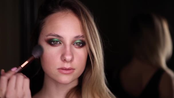 Mooi meisje met groene ogen doet make-up met een borstel op een zwarte achtergrond. Close-up portret — Stockvideo