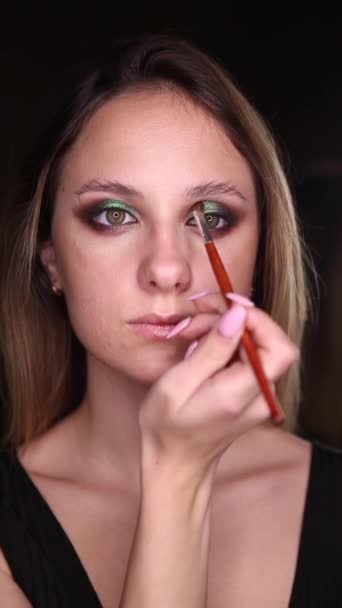 Beautiful girl with green eyes does makeup with a brush on a black background. Vertical video — Stock Video