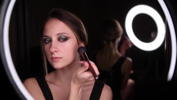 Menina bonita com olhos verdes faz maquiagem com uma escova em um fundo preto. Retrato de close-up — Vídeo de Stock