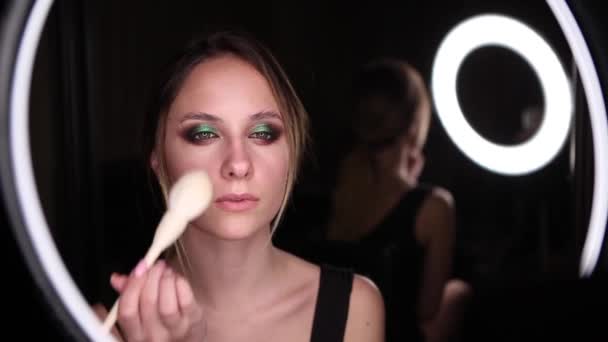 De vrouw doet make-up. Jonge vrouw blush op haar gezicht met een witte borstel make-up. Cosmetisch concept.Zwarte achtergrond, close-up portret — Stockvideo