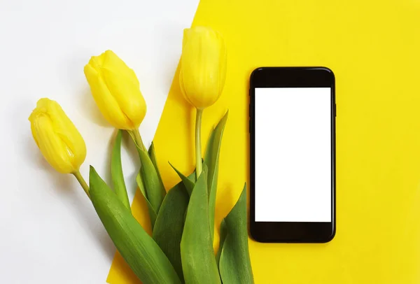 鲜花和一款智能手机在桌子上。黄色的郁金香，黑色智能手机 图库图片
