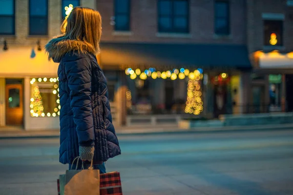 Geceleri alışveriş poşetleri taşıyan bir kadının, mağaza vitrinlerinde Noel doktorları olan bir fotoğrafı.