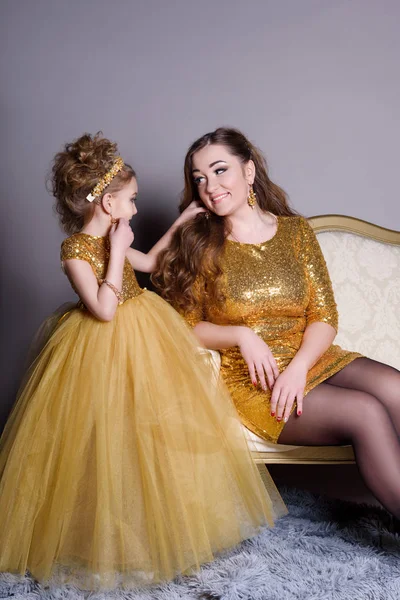 Small beautiful smiling baby with his mother in a smart dress show costume jewelery for yourself on a gray background with a large gold sofa and fluffy gray carpet — Stock Photo, Image