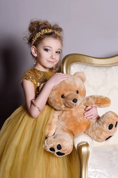 Small beautiful smiling baby in a smart dress jewelry shows on itself on a gray background with a large gold sofa and fluffy gray carpet — Stock Photo, Image