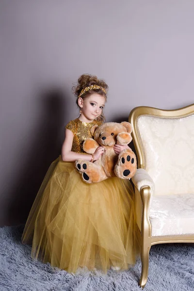 Small beautiful smiling baby in a smart dress jewelry shows on itself on a gray background with a large gold sofa and fluffy gray carpet — Stock Photo, Image