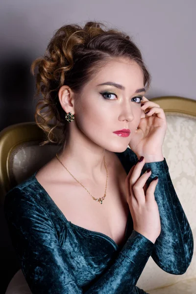 Fată frumoasă într-o rochie de catifea verde pe un fundal gri la frumoasa canapea aurie spectacole de bijuterii — Fotografie, imagine de stoc