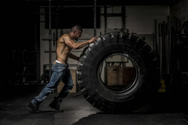 Homem muscular em jeans empurrando pneu grande — Fotografia de Stock