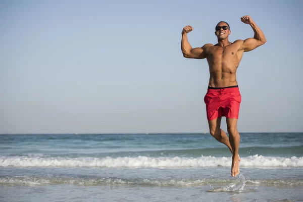 Зрелый пожилой мускулистый мужчина с Ближнего Востока, прыгающий на пляже без рубашки, в красных шортах и солнечных очках — стоковое фото