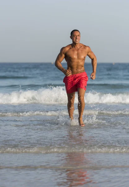 Maduro mais velho muscular ajuste Médio Oriente Árabe Masculino correndo para fora do oceano na praia, sem camisa, vestindo shorts vermelhos — Fotografia de Stock