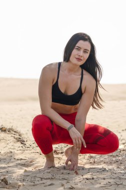 Parlak kırmızı tayt ve siyah spor sutyen giyen güzel esmer fitness modeli, güneşli bir günde sıcak bir Arap çölünde çömelmiş vaziyette duruyor.