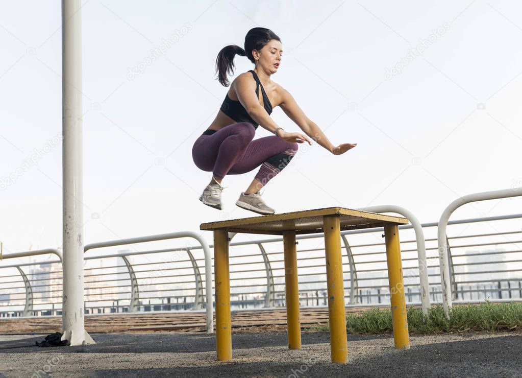 Beautiful brunette fitness model wearing a black sports top and purple tights does a box jump exercise on a bright sunny day.