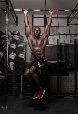 Kaslı, Afrikalı, üstsüz, terli erkek vücut geliştirme sporcusu. Asılı bacak kaldırma hareketi yapıyor. Kaslı, kirli spor salonunda karın kaslarını gösteriyor.