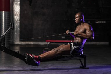Kas yığını Afrikalı Amerikalı üstsüz erkek vücut geliştirme sporcusu. Karanlık, kirli bir spor salonunda, dramatik ışıklandırmayla kürek çekiyor. 