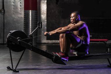 Kas yığını Afrikalı Amerikalı üstsüz erkek vücut geliştirme sporcusu. Karanlık, kirli bir spor salonunda, dramatik ışıklandırmayla kürek çekiyor. 