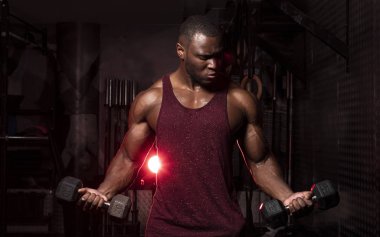 Kas yığını Afrikalı Amerikalı erkek vücut geliştirme sporcusu koyu renkli bir spor salonunda dramatik bir aydınlatma fişeğiyle kıvrımlar yapıyor. 