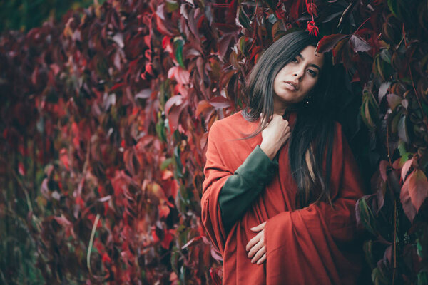 Scared beautiful girl with long natural black hair on background of green red grape hedge. Moody girl in red shawl and green coat hiding in girlish grape. Female emotional portrait in faded tones.
