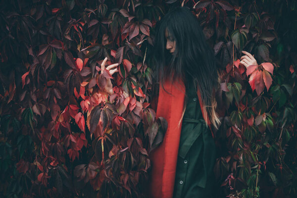 Dreamy beautiful girl with long natural black hair on autumn background of green red grape hedge. Inspired girl in red shawl and green coat. Autumn euphoria. Female emotional portrait in faded tones.