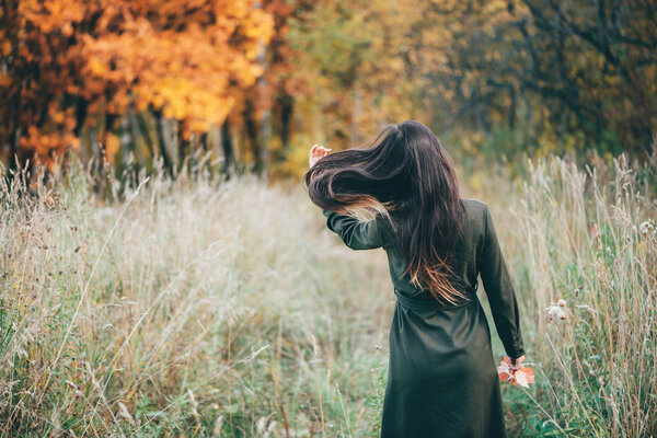 Dreamy beautiful girl with long natural black hair on background with colorful leaves. Fallen leaves in girl hands in autumn forest. Girl surrounded by vivid foliage. Back view. No face.