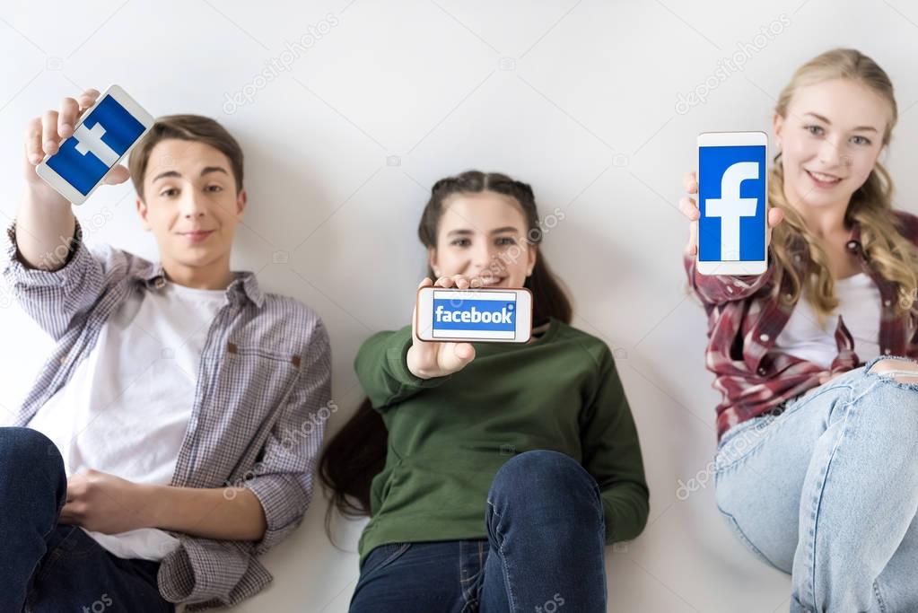 Smiling teenage friends showing smartphones with facebook logo isolated on grey