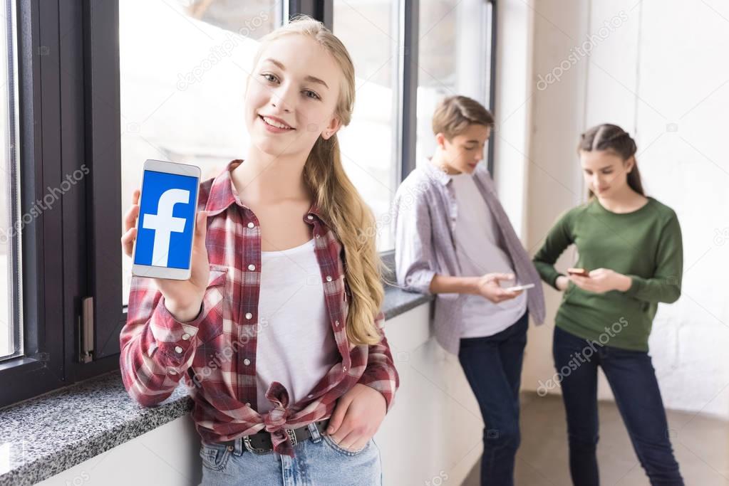 Smiling teenage girl showing smartphone with facebook logo with friends behind