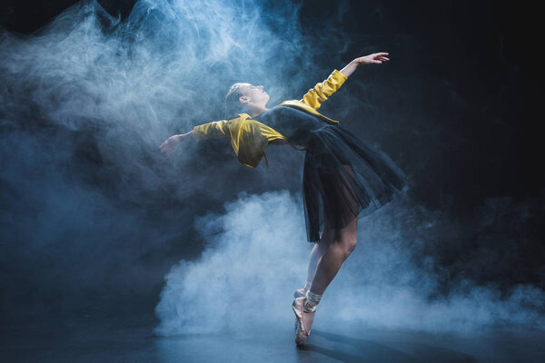 Elegant girl dancing in pointe shoes and black tutu and yellow leather jacket in studio with smoke