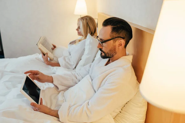 本を読むとデジタル タブレットを使用してホテルの部屋のベッドでバスローブで成熟したカップルの側面図  — 無料ストックフォト