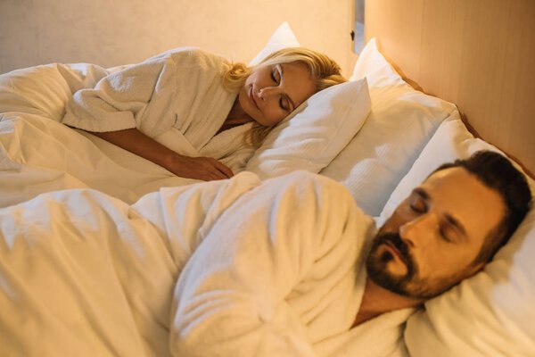 beautiful mature couple in bathrobes sleeping together in bed in hotel room 