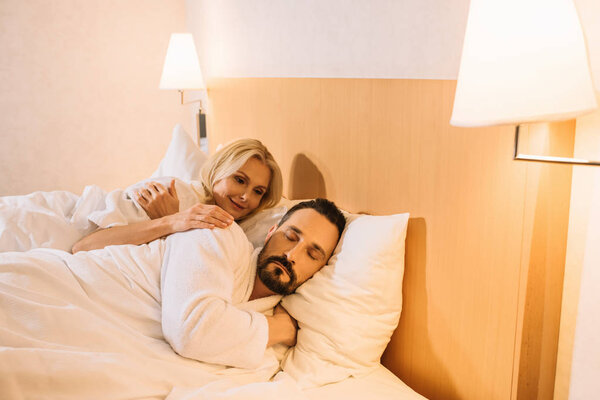 smiling mature woman in bathrobe looking at her husband sleeping in hotel room